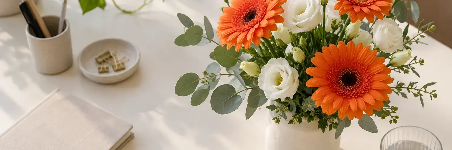 Orange Gerbera und weiße Lisianthus in einer Keramikvase auf einem hellen Schreibtisch.