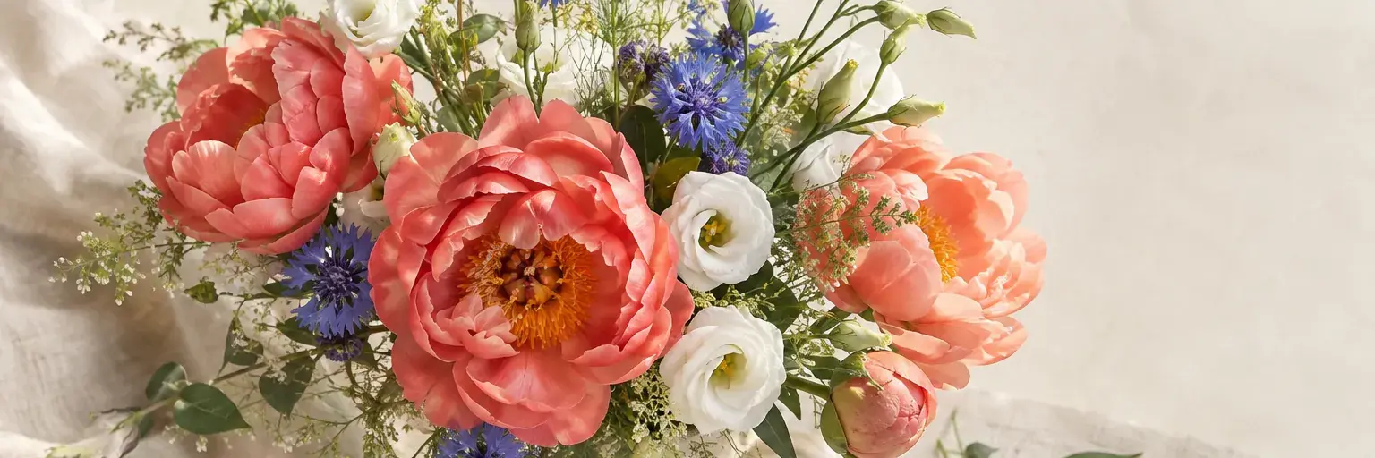 Üppiger Strauß aus korallfarbenen Pfingstrosen, weißen Lisianthus und blauen Kornblumen.
