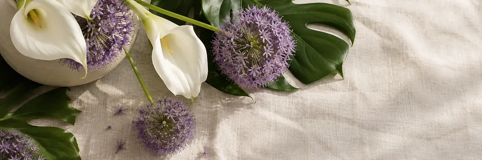 Weiße Calla, violette Zierlauchblüten und grüne Monstera-Blätter in einem schlichten Keramikgefäß.