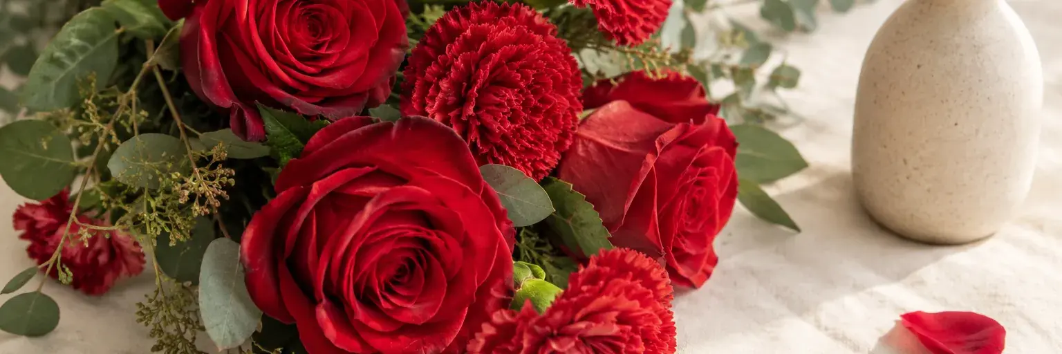 Rote Rosen und Nelken neben einer kleinen Keramikvase.