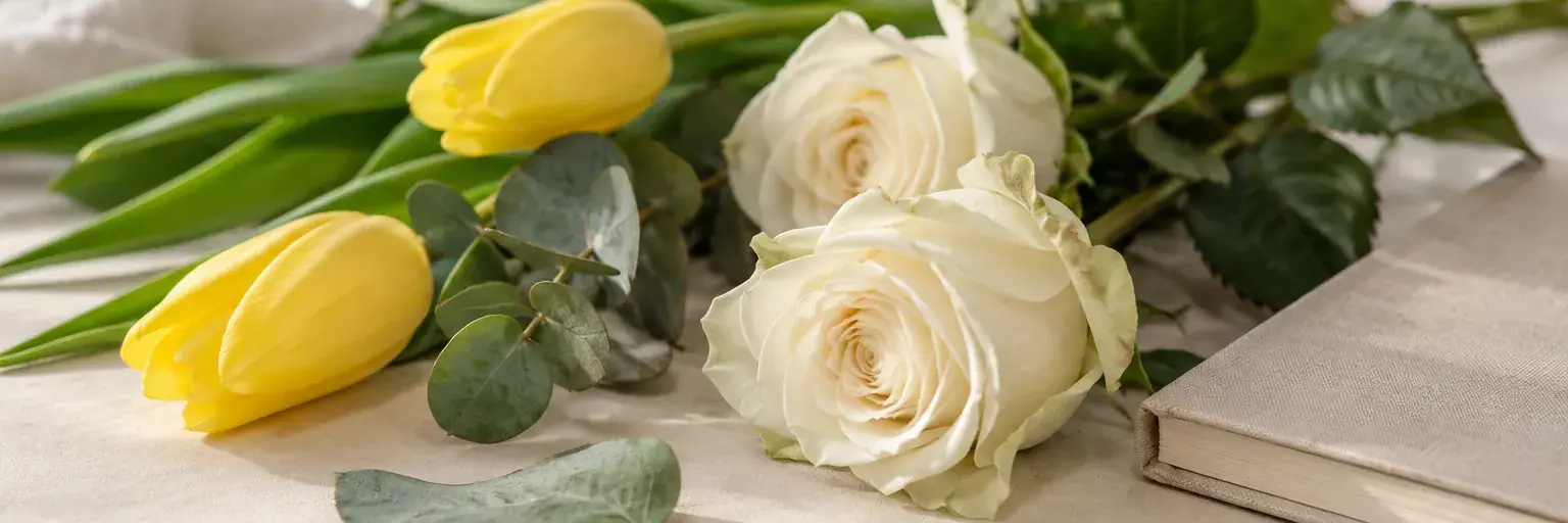 Weiße Rosen und gelbe Tulpen neben einem geschlossenen Notizbuch.