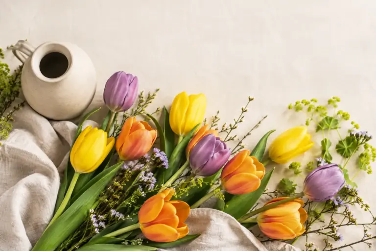Gelbe, orangefarbene und violette Tulpen neben einem schlichten Keramikkrug.